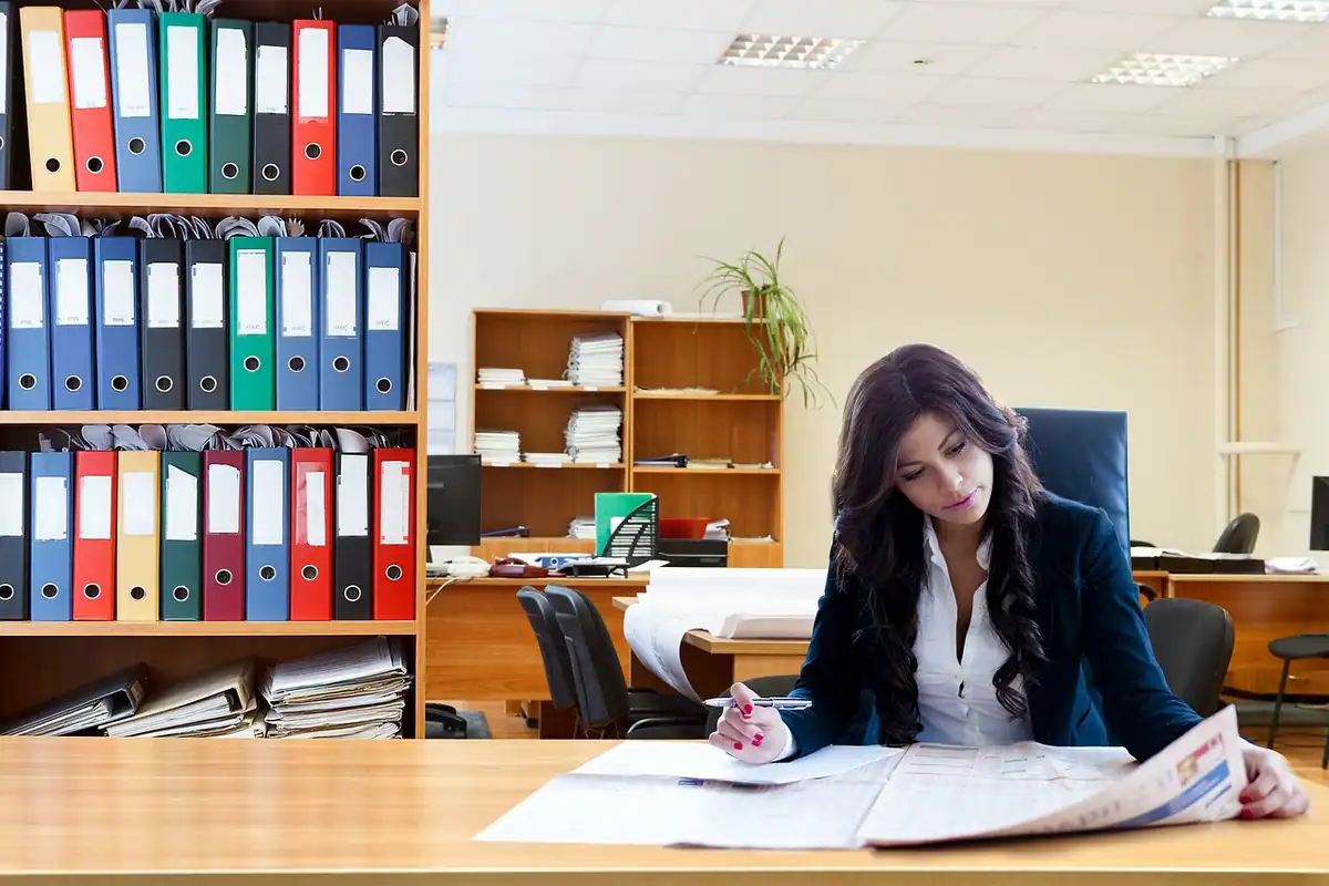 Funcționară la birou analizând documente fiscale, cu bibliorafturi colorate pe rafturi
