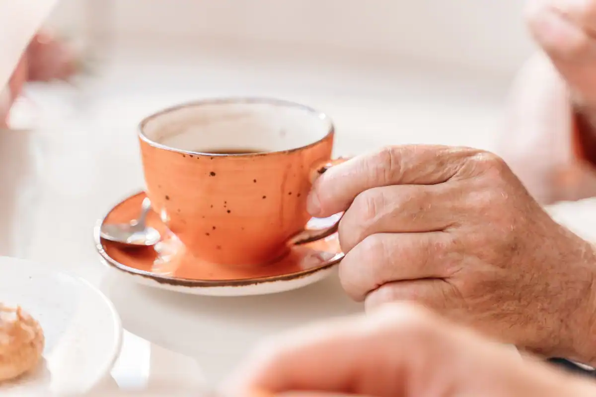 Mână de vârstnic ținând o ceașcă portocalie de cafea, într-o casă caldă