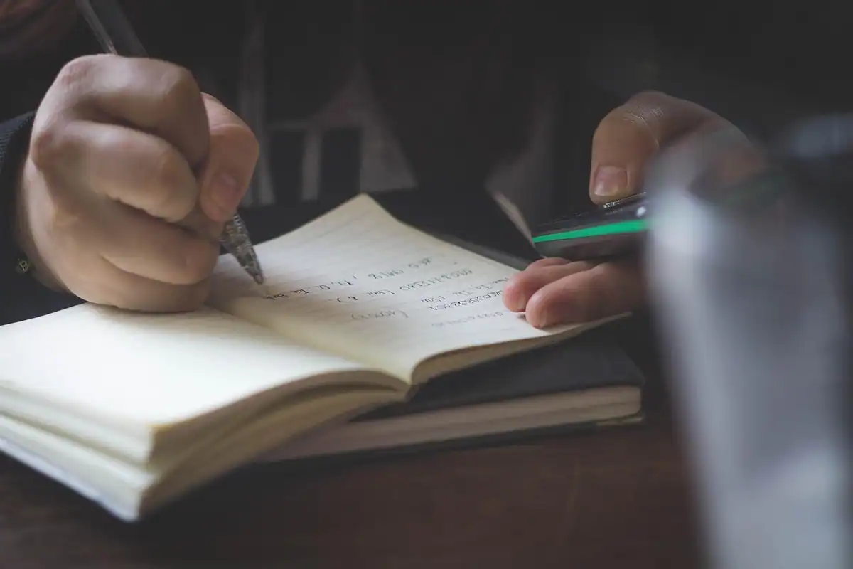 Mână care scrie cu pixul într-un carnet deschis pe o masă întunecată